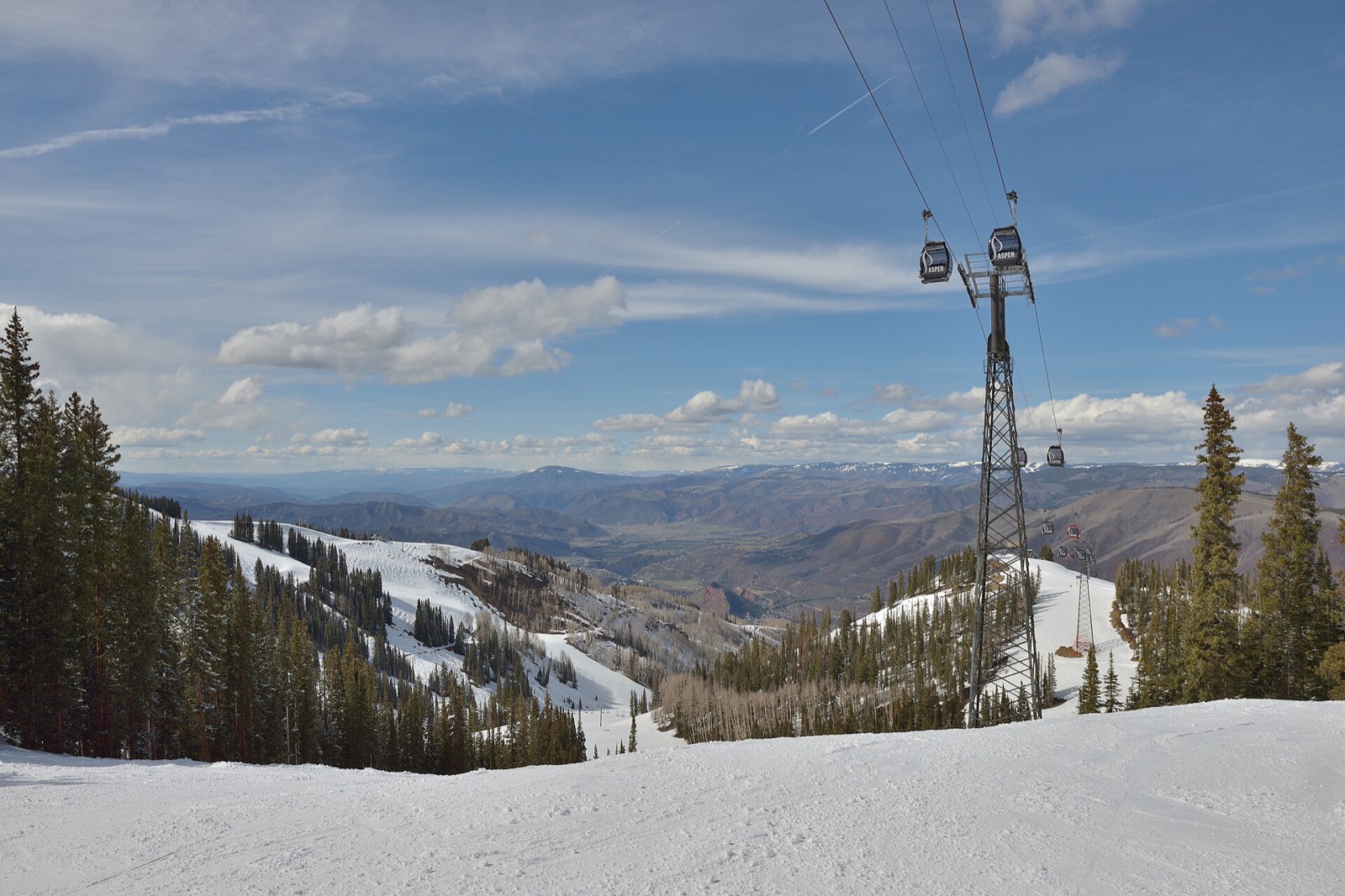 The Silver Queen Gondola crossing a groomed run on Aspen Mountain, with snow-covered conifers and the Roaring Fork valley below