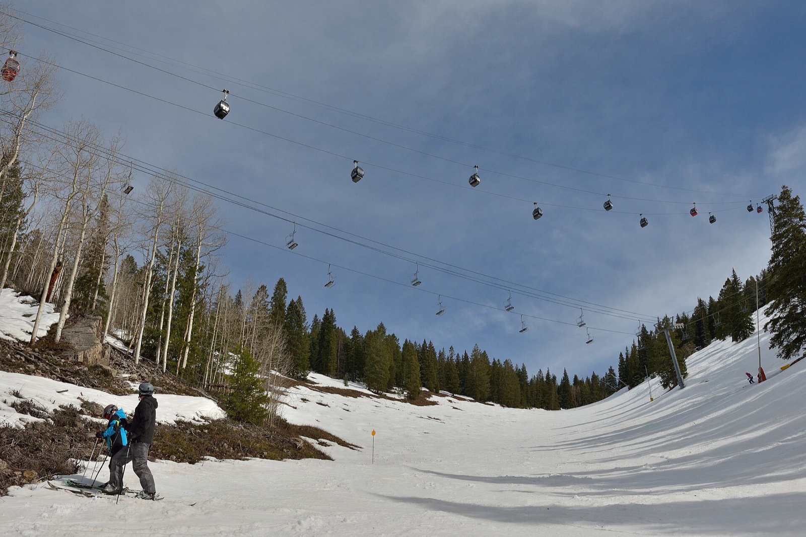 A walker on a bare, thawing slope below the Silver Queen Gondola cabins on Aspen Mountain in spring, with patchy snow remaining